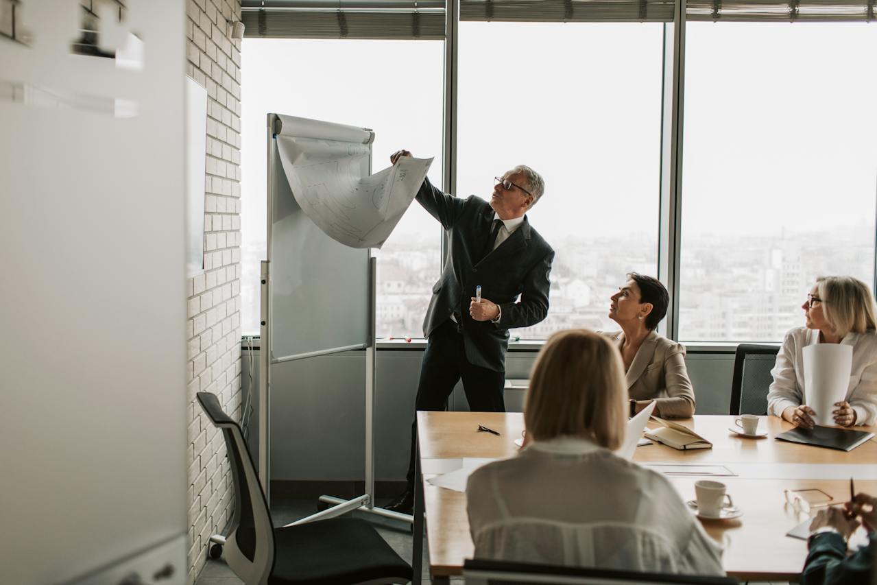 4 people in a meeting room, 3 sitting down, and one holding a marker flipping a page on the board